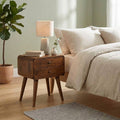 Curved chestnut bedside table with 2 drawers next to a bed with linen bedding